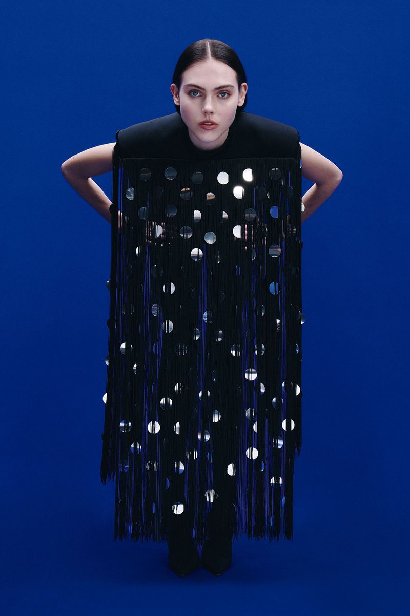 Fashion shoot of a young woman wearing a black dress with reflective circles on a blue background, looking at the camera.