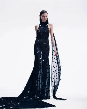 Model in a sheer black halterneck evening dress with metallic polka dots and a long train against a white background