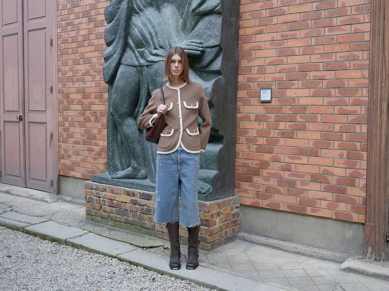 Fashionable adult woman in brown jacket, denim culottes, and boots poses outdoors in front of a statue and brick building.