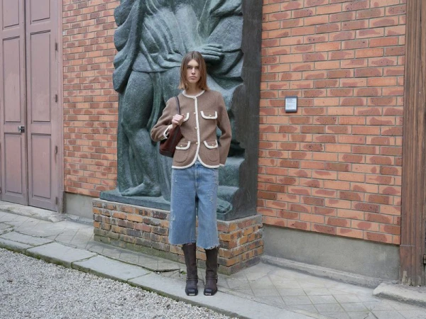 Fashionable adult woman in brown jacket, denim culottes, and boots poses outdoors in front of a statue and brick building.