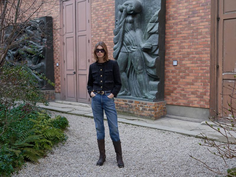 Adult female in tweed jacket, jeans, and boots stands outside by a brick building with sculptural art and lush greenery in daylight.