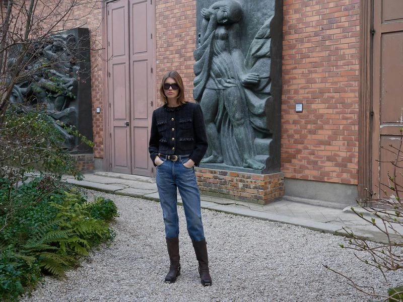 Adult female in tweed jacket, jeans, and boots stands outside by a brick building with sculptural art and lush greenery in daylight.