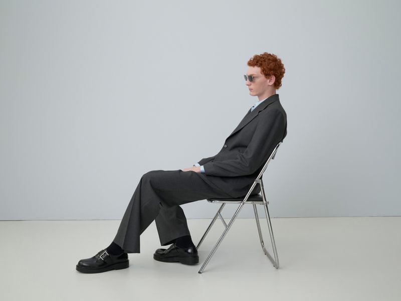 A red-haired man in a grey suit and sunglasses sits in profile on a chrome folding chair against a pale grey studio backdrop. He wears black leather shoes.