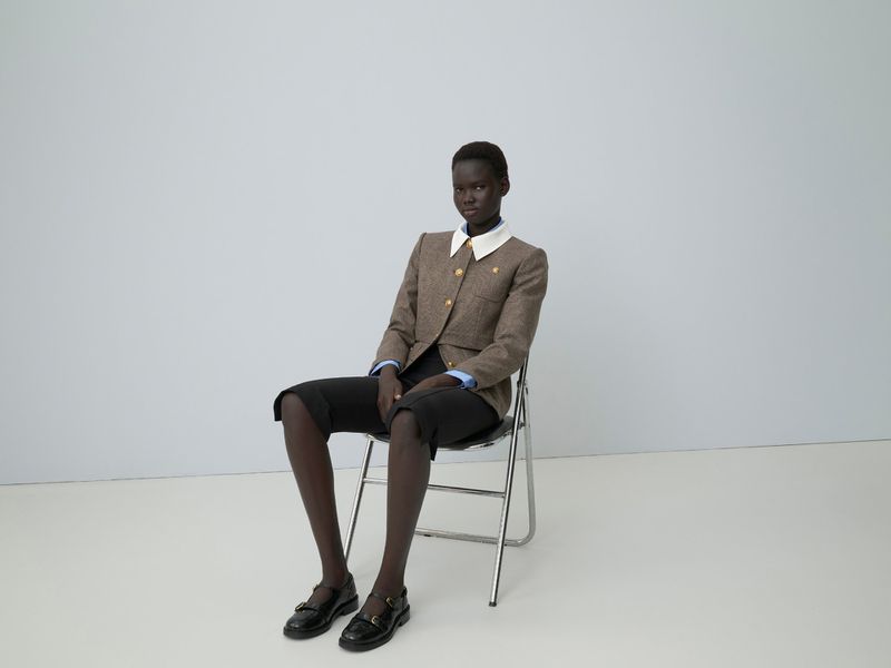 Model seated on a folding chair wearing a brown tweed jacket and dark trousers against a neutral studio backdrop