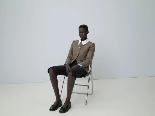 Model seated on a folding chair wearing a brown tweed jacket and dark trousers against a neutral studio backdrop