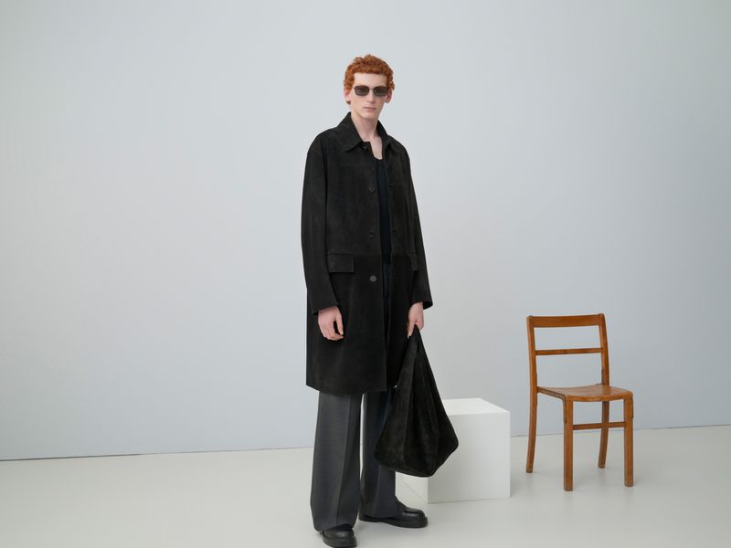 Red-haired male model poses indoors wearing sunglasses, a black overcoat and holding a black bag, next to a wooden chair.