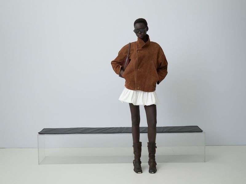 Fashion model poses in a brown jacket, white skirt, and boots in a studio with natural lighting against a white backdrop.