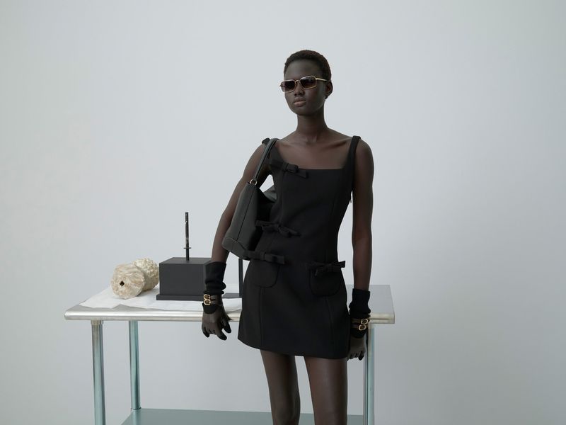 Dark-skinned fashion model in a black mini-dress, carrying a black handbag, wearing sunglasses, standing in a white photo studio.