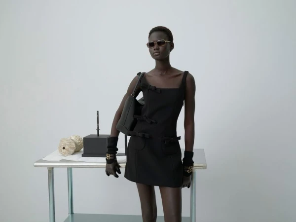 Dark-skinned fashion model in a black mini-dress, carrying a black handbag, wearing sunglasses, standing in a white photo studio.