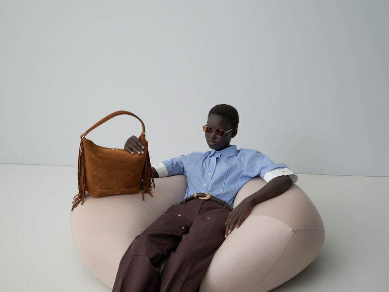 Model wearing a blue button-down shirt and brown trousers seated in a cream pod chair holding a brown fringed suede bag against a grey seamless backdrop