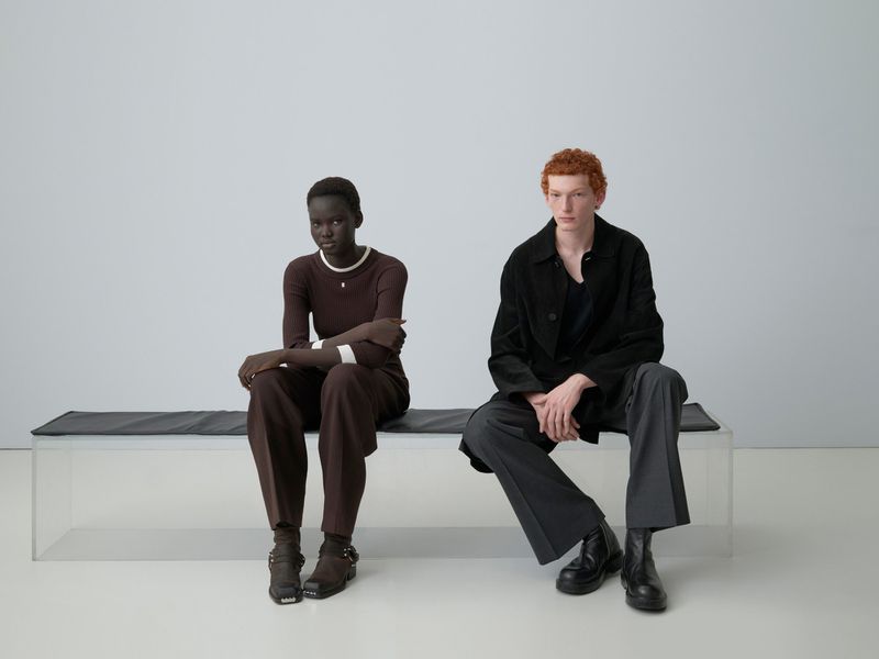 Two fashion models, an African woman and a Caucasian man with red hair, sit on a bench in a minimalist photo studio setting.