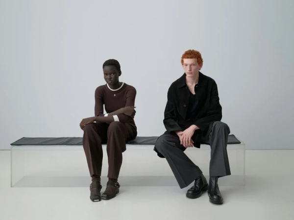 Two fashion models, an African woman and a Caucasian man with red hair, sit on a bench in a minimalist photo studio setting.