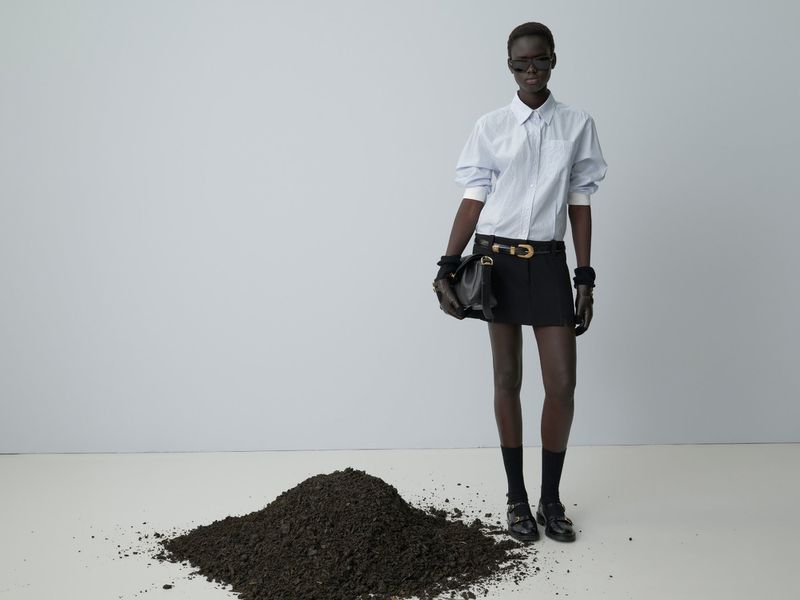 Editorial fashion portrait of a Black woman in a striped shirt and skirt standing on a white floor with a pile of dirt, studio shot.