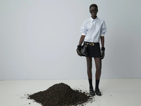Editorial fashion portrait of a Black woman in a striped shirt and skirt standing on a white floor with a pile of dirt, studio shot.