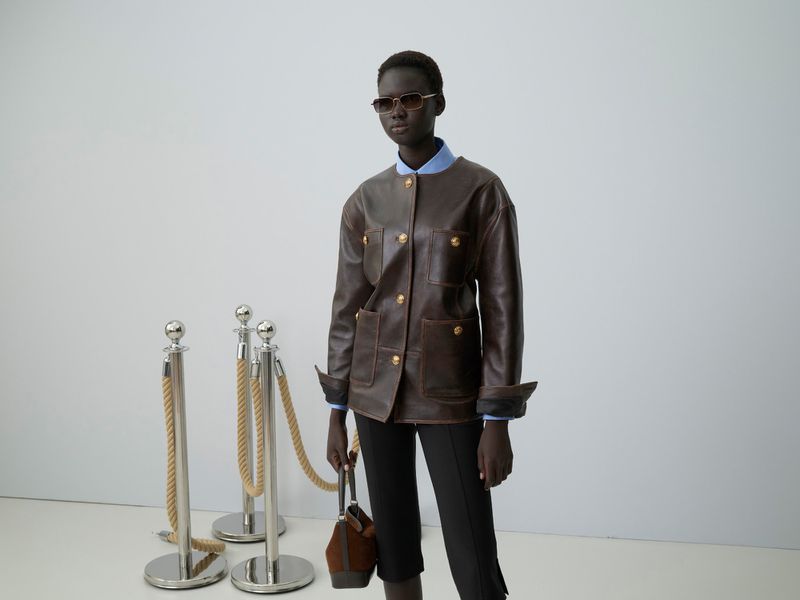 Dark-skinned woman with sunglasses, brown leather jacket, and black pants stands in front of silver stanchions with rope.