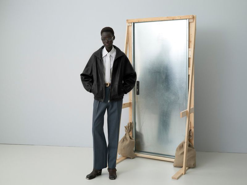Fashion model posing in a black jacket and grey pants in front of a metal door with burlap sacks. Grey background, indoor setting.