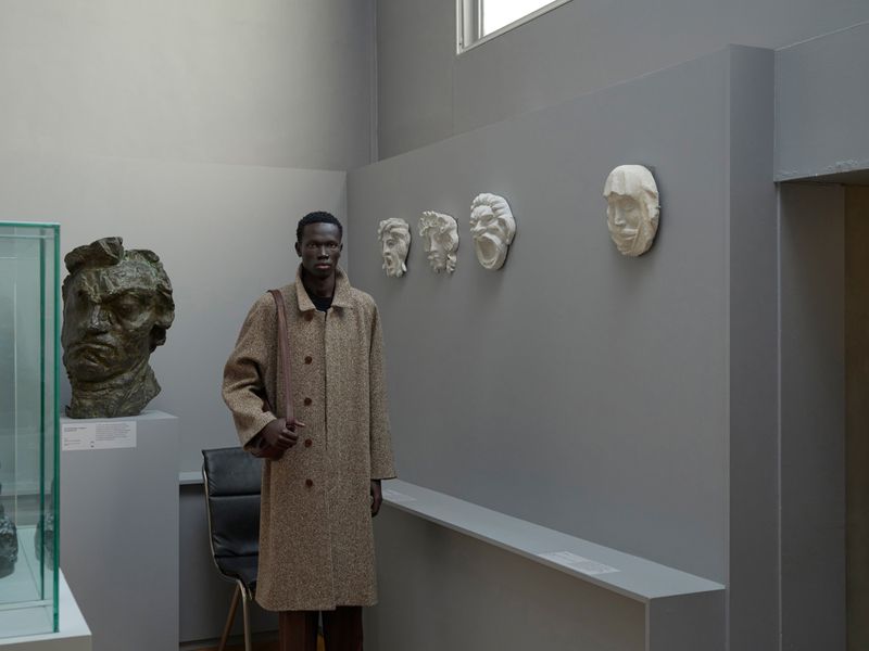 A dark-skinned man stands in an art gallery next to sculptures. He wears a brown overcoat, carrying a small bag, with art on the wall.