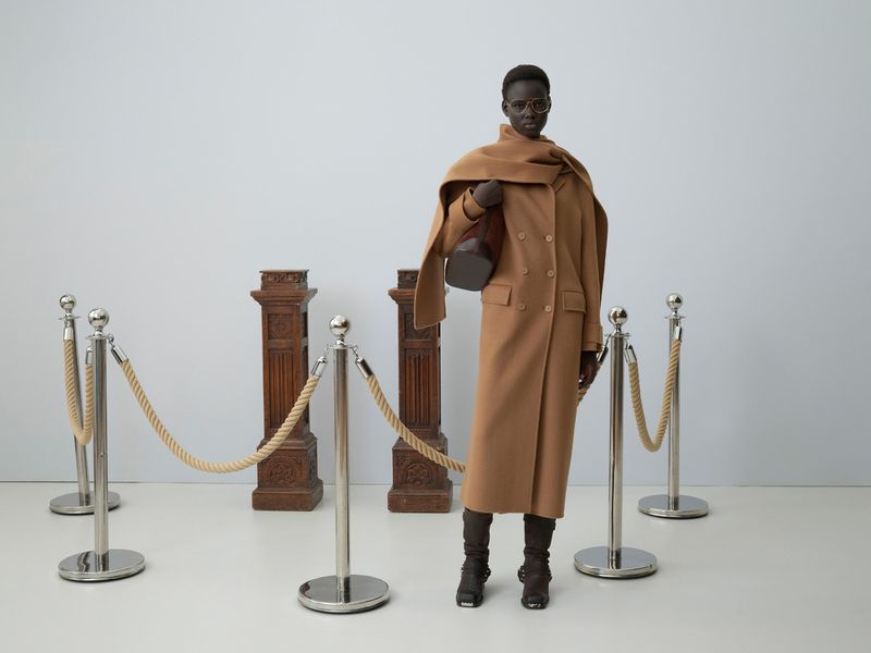 Woman in a tan coat and glasses, holding a brown purse, poses indoors with rope barriers and decorative pillar-like objects in the background.