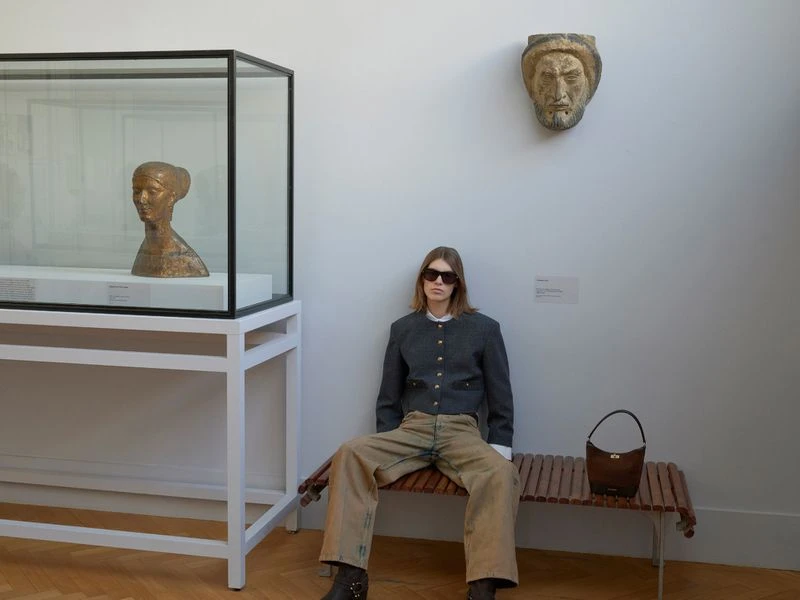 Woman in sunglasses sits on a bench in an art museum next to a display case with a bust and a wall-mounted sculpture.