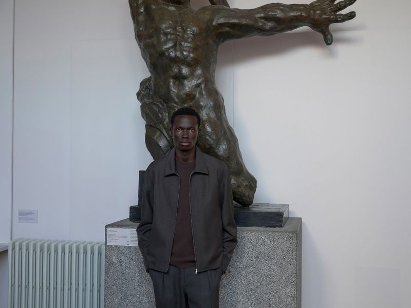 Portrait of an African American man in a jacket standing indoors in front of a bronze statue in a museum setting on a gray background.