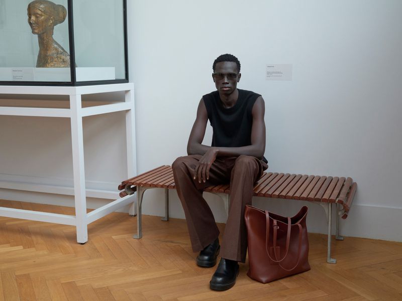 An adult Black man sits on a wood and metal bench next to a brown tote bag indoors with a statue display case in the background.