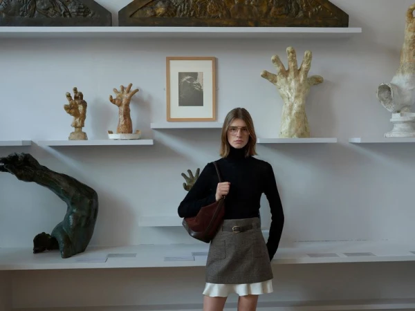 Model wearing a black turtleneck and plaid skirt holding a brown shoulder bag inside a gallery with sculptures