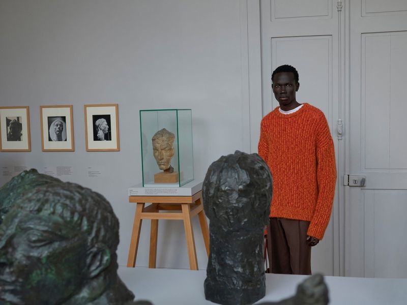 In a museum setting, an adult male model in an orange sweater stands next to classical busts and framed portraits on the wall.