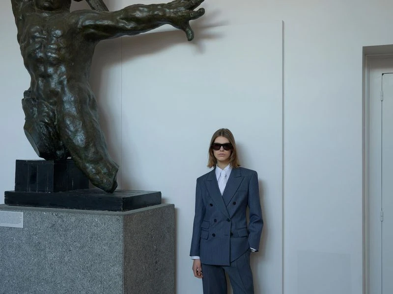 Woman in pinstripe suit poses indoors beside a classical statue. She wears sunglasses and a white collared shirt under the grey jacket.