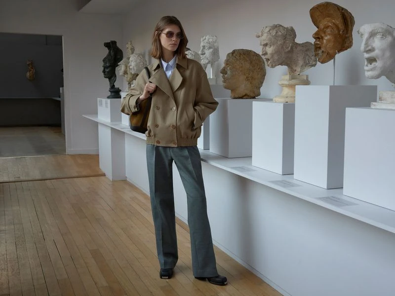 Woman in a tan jacket and gray pants stands in a gallery with classical bust sculptures on display and light wood flooring.