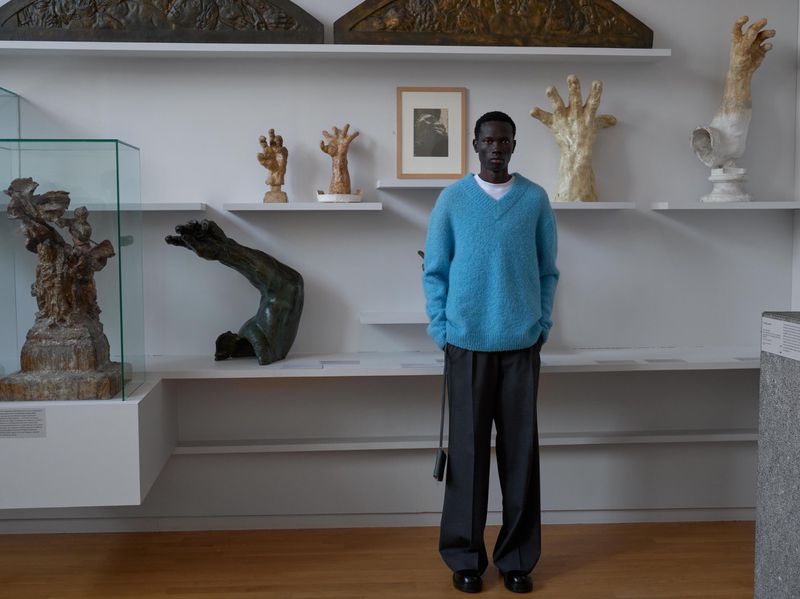 Man stands in an art gallery wearing a blue sweater and gray pants, surrounded by shelves of sculptures and art pieces.