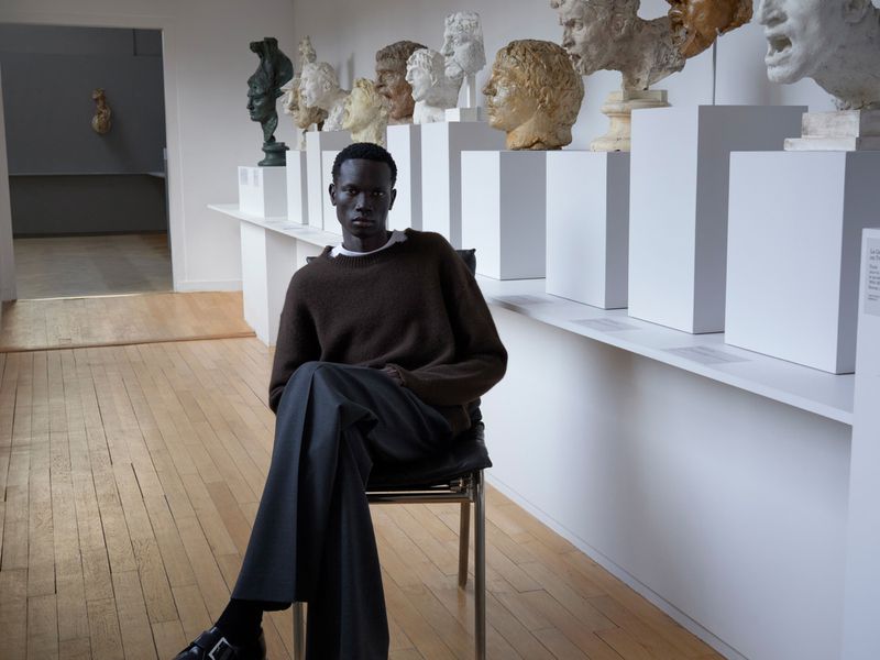 Dark-skinned male fashion model poses in a brown sweater and grey slacks on a chair in a light-filled art gallery with busts.