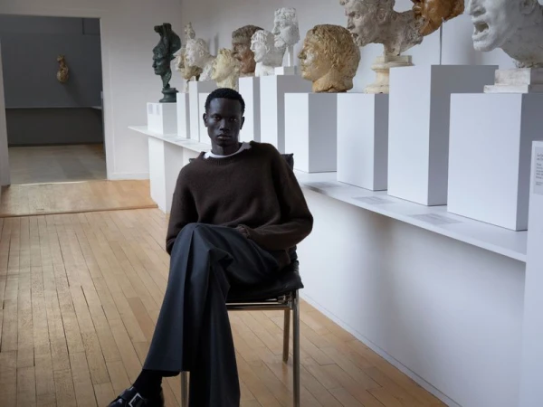 Dark-skinned male fashion model poses in a brown sweater and grey slacks on a chair in a light-filled art gallery with busts.