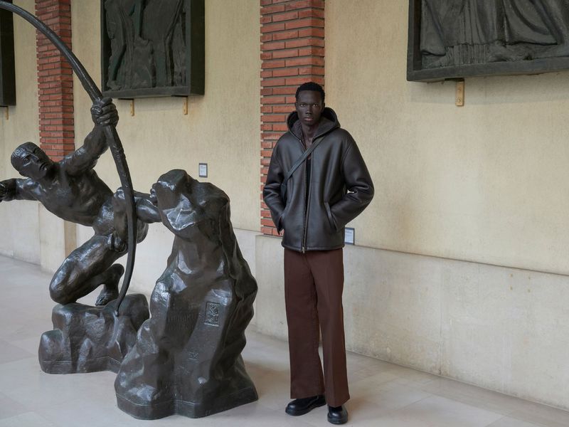 Fashionable adult male in hooded jacket poses next to bronze sculpture in an indoor location with brick architectural details. 