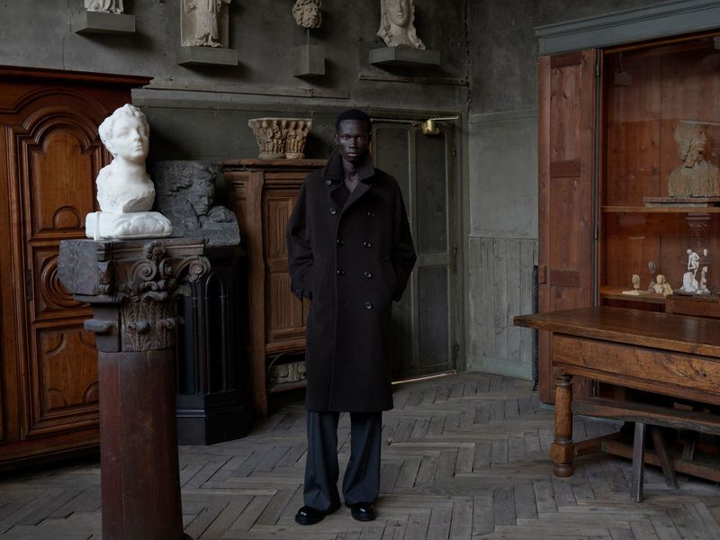 A stylish African American man in a dark brown coat and grey pants stands in a room filled with sculptures and wooden furniture.