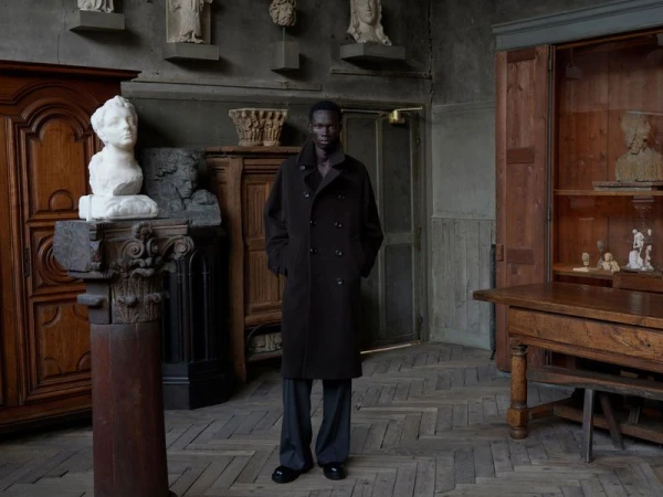 A stylish African American man in a dark brown coat and grey pants stands in a room filled with sculptures and wooden furniture.