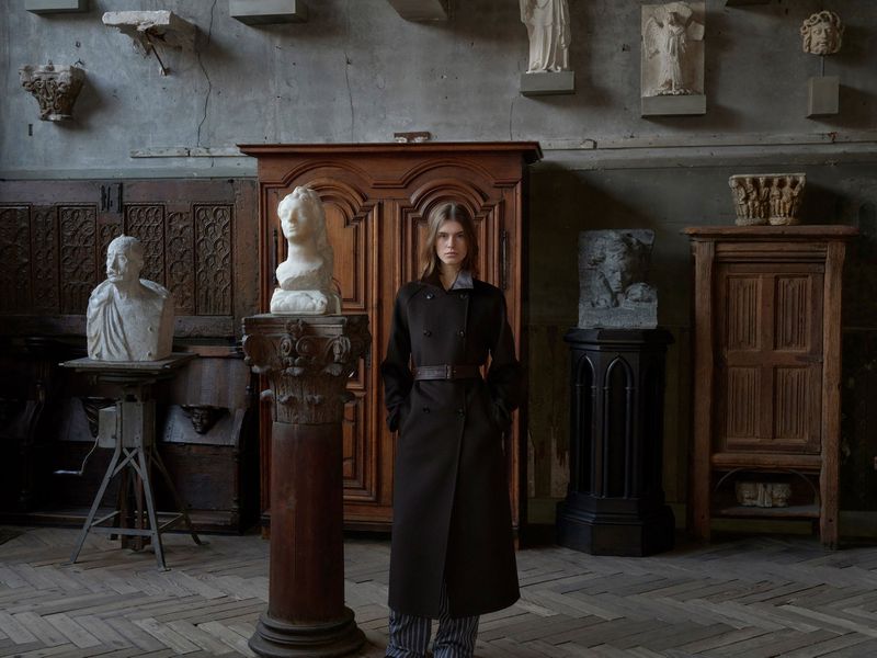 A woman in a dark coat poses indoors amidst sculptures and antique furniture, complemented by a herringbone floor and a subdued, elegant ambiance.