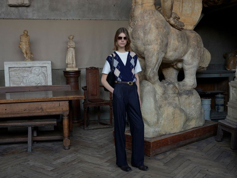 Indoor fashion shot of a young adult woman in a navy sweater vest, pleated trousers, and black sunglasses, posing near a stone sculpture.