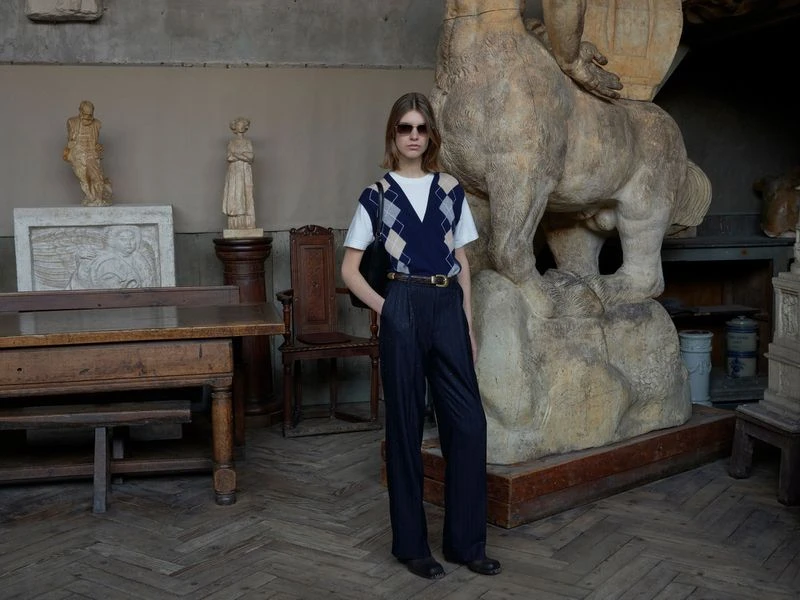 Indoor fashion shot of a young adult woman in a navy sweater vest, pleated trousers, and black sunglasses, posing near a stone sculpture.