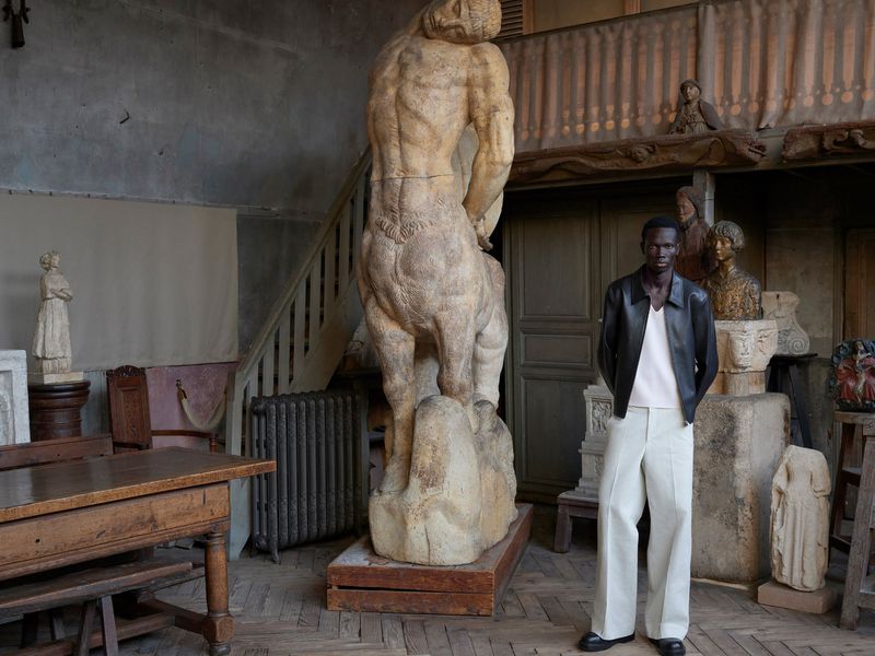 A man posing in front of a centaur statue in an antique building, wearing a black jacket and light-colored pants.