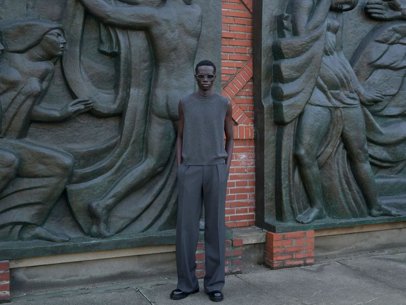 An adult Black male model in a sleeveless sweater and trousers stands in front of an artistic stone relief on a brick and stone base.