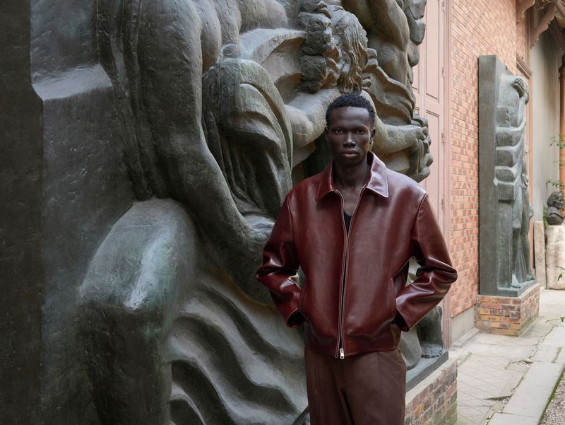 An adult male poses wearing a leather jacket in front of carved stone artwork and a brick wall outdoors during daytime.