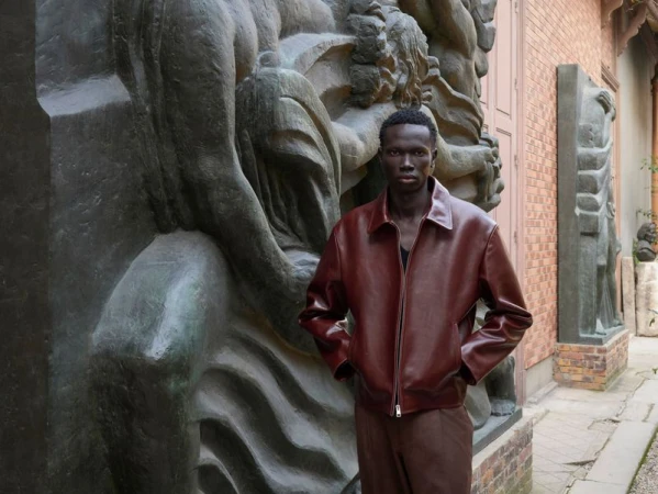 An adult male poses wearing a leather jacket in front of carved stone artwork and a brick wall outdoors during daytime.