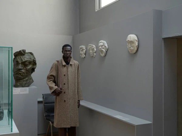 Man wearing a brown textured coat standing in a museum gallery next to sculpted busts and plaster face casts on a grey wall.