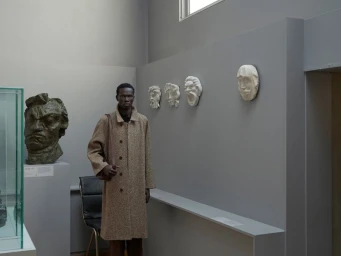 Man wearing a brown textured coat standing in a museum gallery next to sculpted busts and plaster face casts on a grey wall.