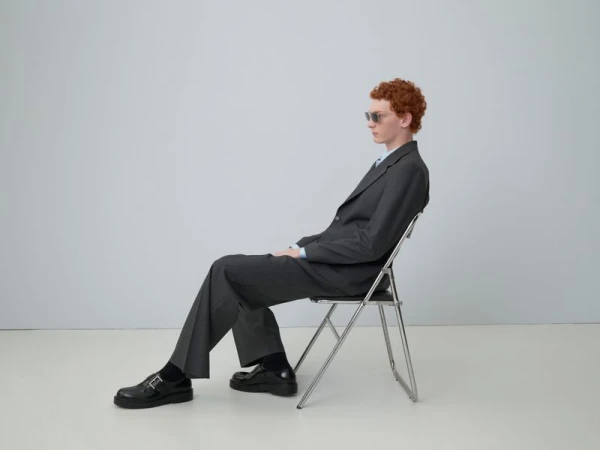 Man wearing a dark grey suit sitting on a metal folding chair against a neutral grey studio background