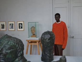 Model wearing an orange ribbed sweater in a gallery setting with classical sculptures and framed artworks on the wall.
