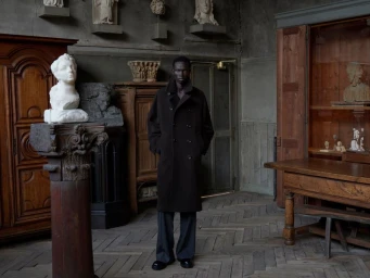 Man wearing a long dark coat standing in an interior space filled with wooden cabinets and classical stone sculptures.