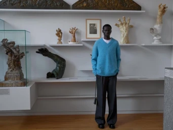 Man wearing a blue V-neck sweater and dark trousers standing inside a museum gallery featuring sculptural art.