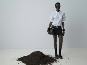 Model standing next to a pile of soil wearing a light blue button-down shirt and black skirt against a grey background.