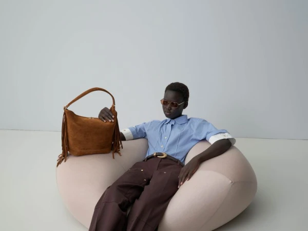Model wearing a blue button-down shirt and brown trousers seated in a cream pod chair holding a brown fringed suede bag against a grey seamless backdrop